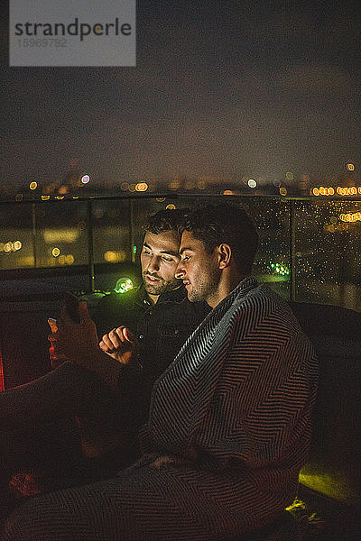 Mann benutzt Smartphone  während er nachts neben einem Freund auf der Terrasse sitzt