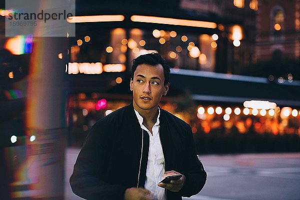 Junger Mann mit Telefon schaut weg  während er nachts in einer beleuchteten Stadt steht