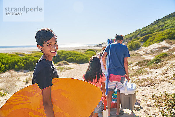 Porträt eines glücklichen Jungen mit Bodyboard am sonnigen Strand mit Familie