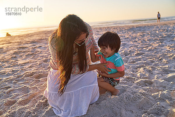 Verspielte Mutter kitzelt Sohn am Strand bei Sonnenuntergang