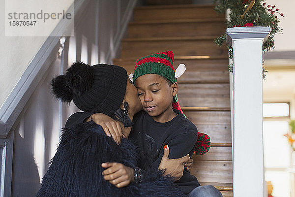Zärtliche Mutter küsst Sohn mit Weihnachtshut auf Treppe