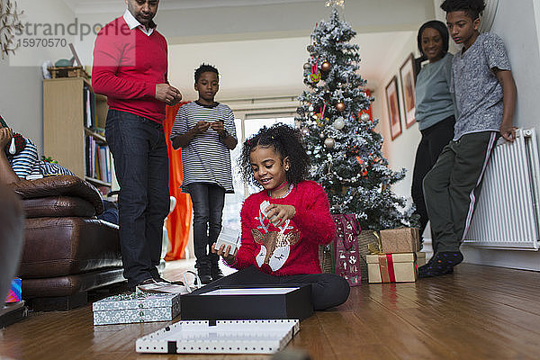 Familie beobachtet Mädchen beim Öffnen von Weihnachtsgeschenken auf dem Wohnzimmerboden