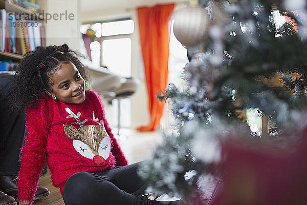 Neugieriges Mädchen im Weihnachtspullover schaut auf Weihnachtsbaum