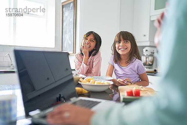 Glückliche Schwestern frühstücken in der Küche  während die Mutter am Laptop arbeitet