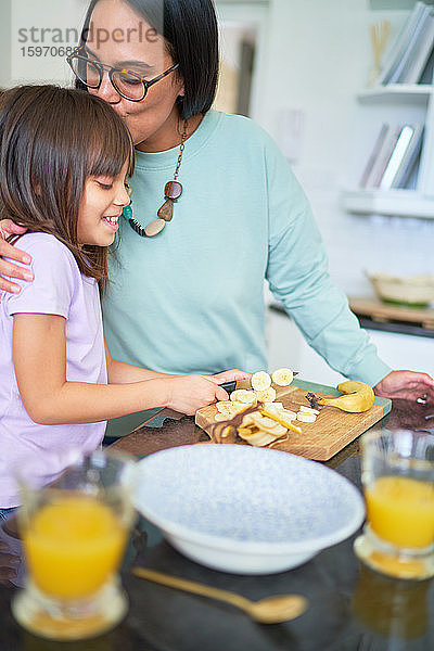 Liebevolle Mutter küsst Tochter  die in der Küche Banane schneidet