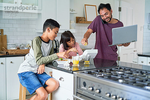 Vater mit Laptop füttert Kinder mit Essen zum Mitnehmen in der Küche