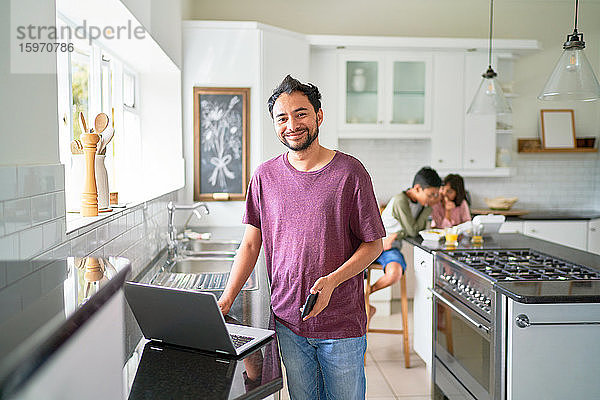 Porträt eines glücklichen Vaters  der am Laptop in der Küche arbeitet  während die Kinder essen