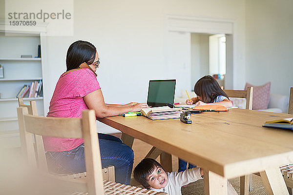 Mutter arbeitet am Laptop mit spielenden Kindern und macht Hausaufgaben