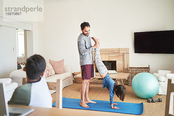 Vater hilft Tochter beim Handstand auf Yogamatte im Wohnzimmer