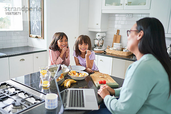 Glückliche Töchter  die frühstücken und der Mutter bei der Arbeit in der Küche zusehen