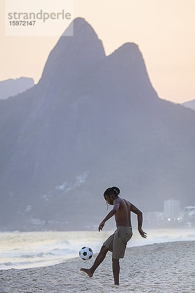 Ein junger schwarzer Brasilianer spielt Fussball am Strand von Ipanema mit dem Dois Irmaos-Gebirge in der Ferne  Rio de Janeiro  Brasilien  Südamerika