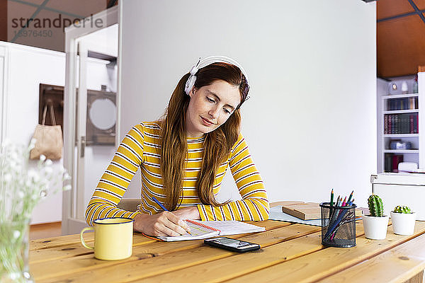 Frau mit Kopfhörern  die in ein Buch schreibt und dabei auf ihr Smartphone auf dem Schreibtisch schaut
