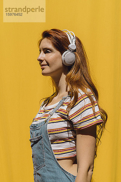 Lächelnde junge Frau hört Musik über Kopfhörer an einer gelben Wand an einem sonnigen Tag
