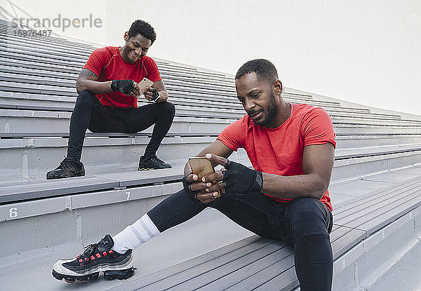 Zwei Sportler sitzen auf einer Treppe in einem Stadion und benutzen Smartphones