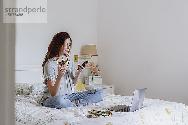 Schöne Frau  die ein Mobiltelefon benutzt  während sie mit Pizza und Laptop auf dem Bett zu Hause sitzt