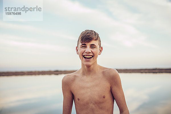 Porträt eines glücklichen Teenagers ohne Hemd  der mit einem See und einem bewölkten Himmel im Hintergrund steht