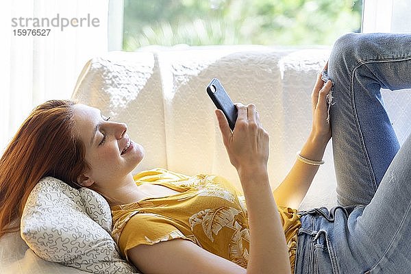 Junge Frau liegt zu Hause auf der Couch und benutzt ihr Smartphone