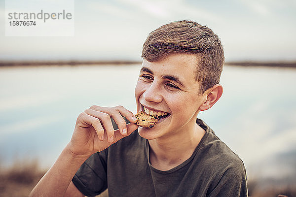 Nahaufnahme eines fröhlichen Teenagers  der einen Keks isst  mit See und Himmel im Hintergrund