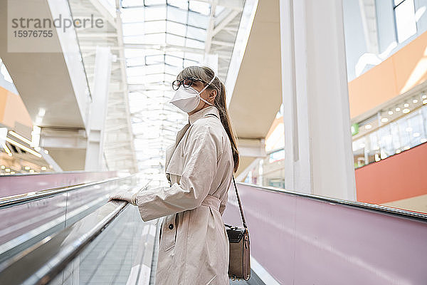 Frau mit Gesichtsmaske und Einweghandschuhen auf einer Rolltreppe in einem Einkaufszentrum