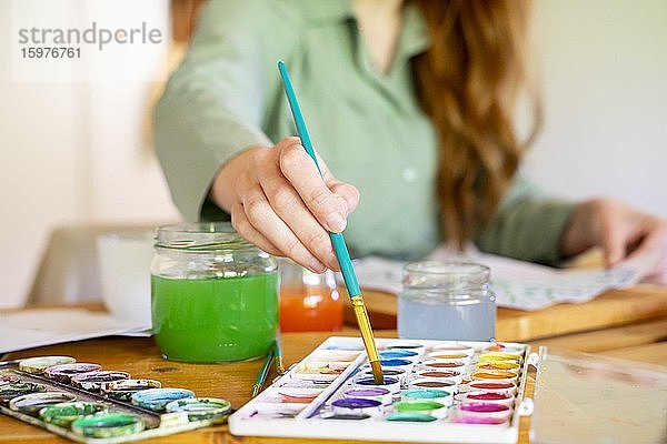 Junge Frau taucht Pinsel in Aquarellfarbe auf einem Tisch zu Hause