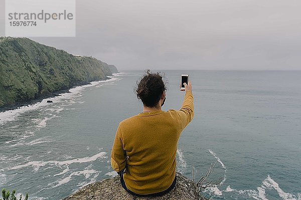 Rückansicht eines Mannes  der auf einem Felsen an der Küste sitzt und ein Smartphone-Foto auf der Insel Sao Miguel  Azoren  Portugal  macht