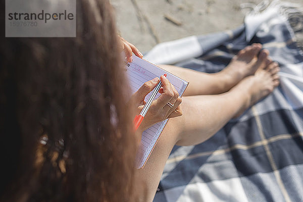 Ansicht einer jungen Frau  die auf einer Decke am Strand sitzt und in ein Notizbuch schreibt