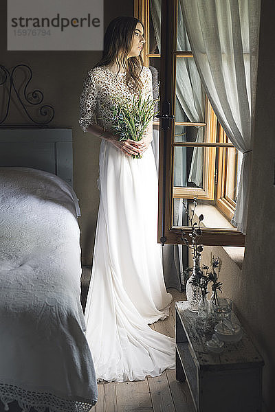 Junge Frau in elegantem Hochzeitskleid  die einen Blumenstrauß hält und aus dem Fenster schaut