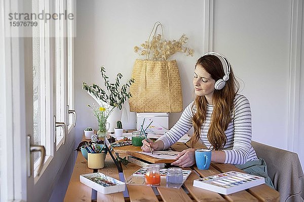 Junge Frau hört Musik über Kopfhörer  während sie zu Hause am Tisch malt