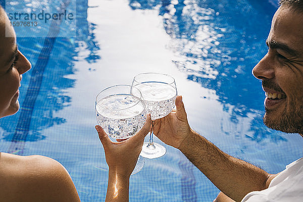 Fröhliches junges Paar  das sich beim Anstoßen mit Getränken am Swimmingpool ansieht
