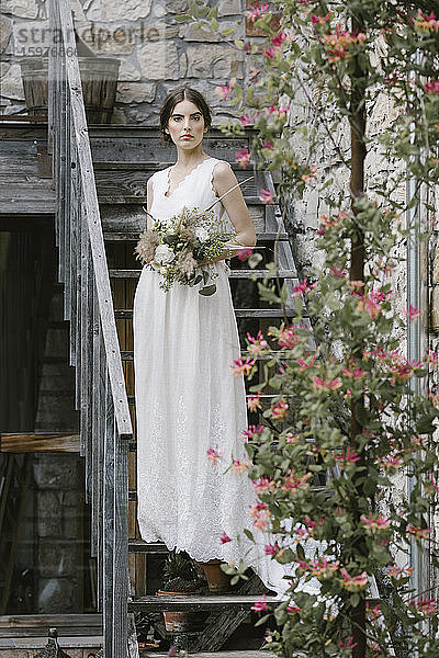 Junge Frau in elegantem Hochzeitskleid mit Blumenstrauß auf einer Treppe stehend