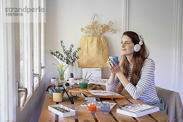 Nachdenkliche junge Frau hält Kaffeetasse und hört Musik  während sie zu Hause auf dem Tisch malt