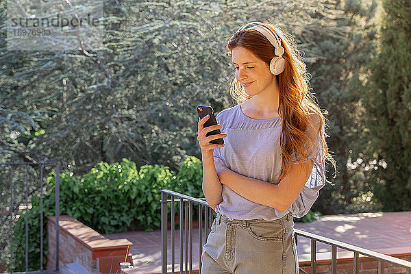 Lächelnde rothaarige Frau mit Kopfhörern und Smartphone auf der Terrasse