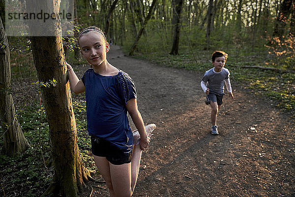Mädchen schaut weg  während sie ihr Bein am Baumstamm gegen den im Wald laufenden Bruder streckt