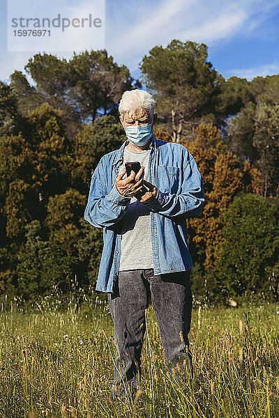 Älterer Mann mit Maske und Smartphone auf einer Wiese stehend