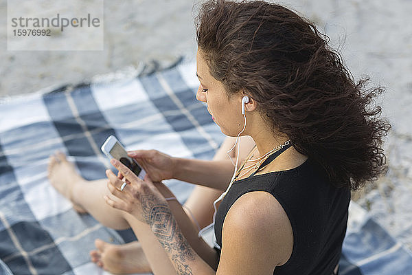 Junge Frau sitzt auf einer Decke am Strand und benutzt Kopfhörer und Smartphone