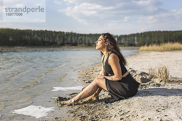 Junge Frau sitzt am Seeufer und genießt das Sonnenlicht