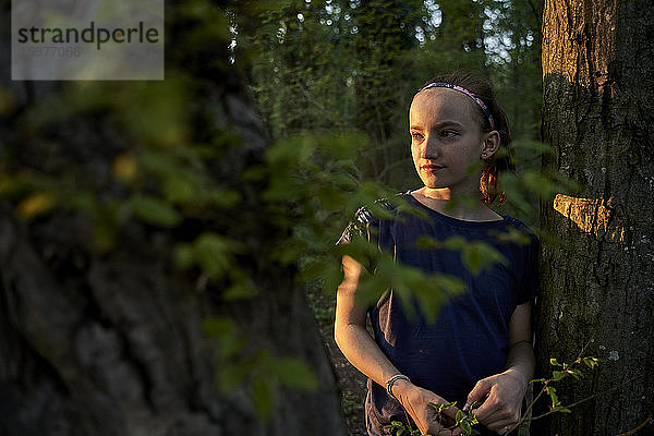 Mädchen steht an einem Baumstamm und schaut im Wald weg
