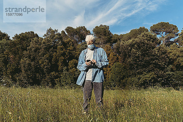 Älterer Mann mit Maske und Smartphone auf einer Wiese stehend