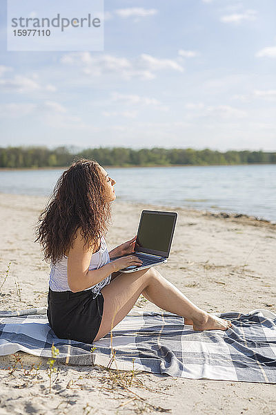 Junge Frau sitzt auf einer Decke am Strand und benutzt einen Laptop