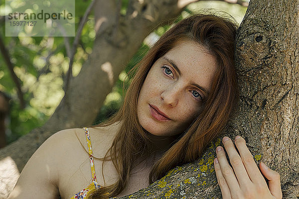 Nachdenkliche junge Frau  die sich an einen Baumstamm im Garten lehnt und den Blick abwendet
