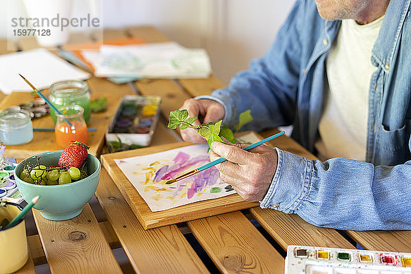 Älterer Mann hält Blätter beim Malen auf dem Tisch zu Hause