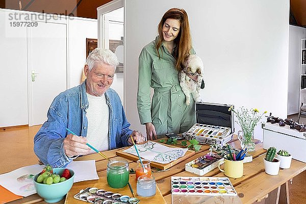 Glückliche Tochter  die eine Katze trägt und einen älteren Mann betrachtet  der auf Papier malt