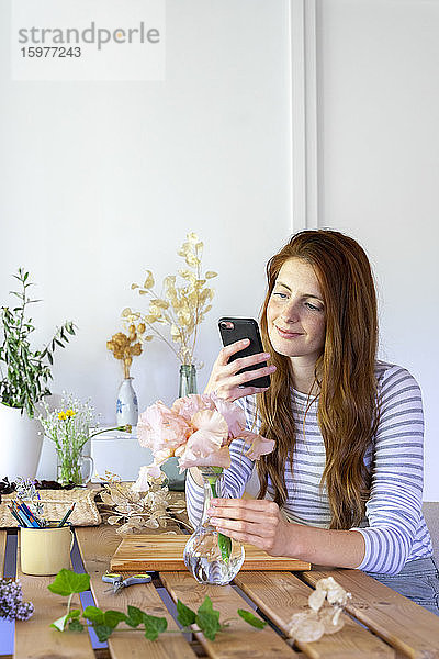 Spanien  Porträt einer jungen  schönen  rothaarigen Frau  die ihr Smartphone benutzt  während sie am Tisch Pflanzen eintopft
