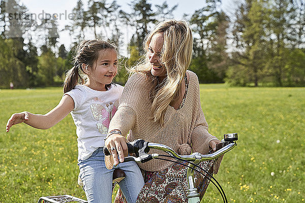 Lächelnde blonde Frau sieht ihre Tochter an  die mit ausgestreckten Armen Fahrrad fährt