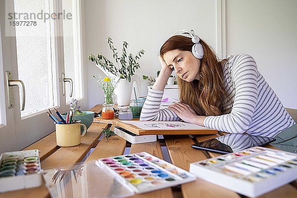 Nachdenkliche Frau hört Musik über Kopfhörer  während sie zu Hause malt