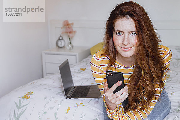 Schöne Frau mit grauen Augen und langen braunen Haaren sitzt mit Handy auf Schlafzimmer zu Hause