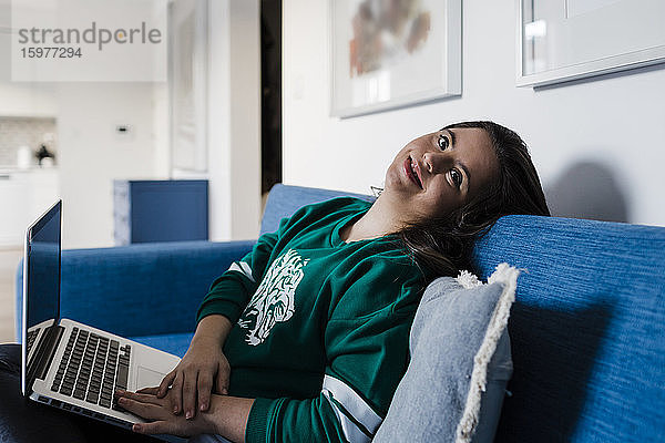 Glückliche junge Frau mit Down-Syndrom benutzt einen Laptop  während sie sich zu Hause auf dem Sofa entspannt