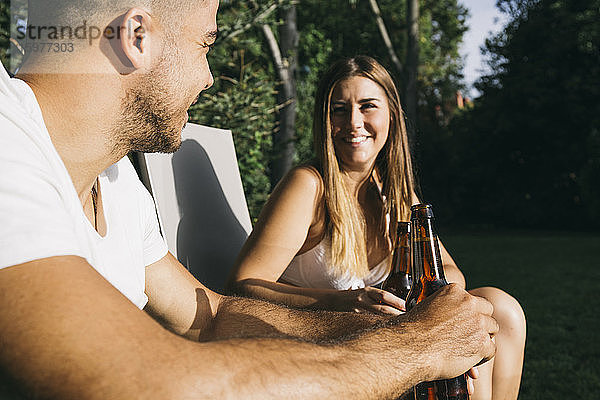 Lächelndes junges Paar stößt mit Bierflaschen an  während es in einem Touristenort sitzt