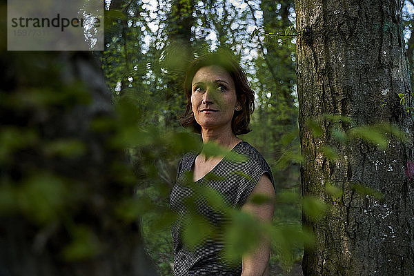 Frau schaut weg  während sie gegen einen Baumstamm im Wald steht