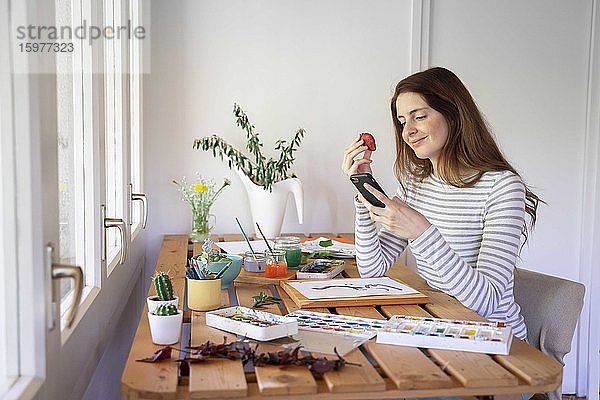 Junge Frau isst eine Erdbeere und benutzt ihr Smartphone  während sie zu Hause auf dem Tisch malt